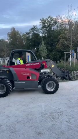 A red telehandler with a person wearing a high-visibility vest driving it moves along a gravel path. The telehandler has large tires and forks extended forward. The background consists of trees and a cloudy sky, suggesting it is either dawn or dusk. Several other vehicles and bicycles are visible in the distance, along with some fencing and structures. The telehandler turns to the right and continues moving down the path.
