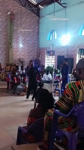 Inside a church, a gospel choir performs on a stage for an audience. The choir consists of drummers, a keyboard player, and singers with microphones. The church is decorated with brick walls, blue arched windows, and ceiling fans. The audience members are seated on blue plastic chairs. A man in a colorful shirt is in the foreground, partially blocking the view.