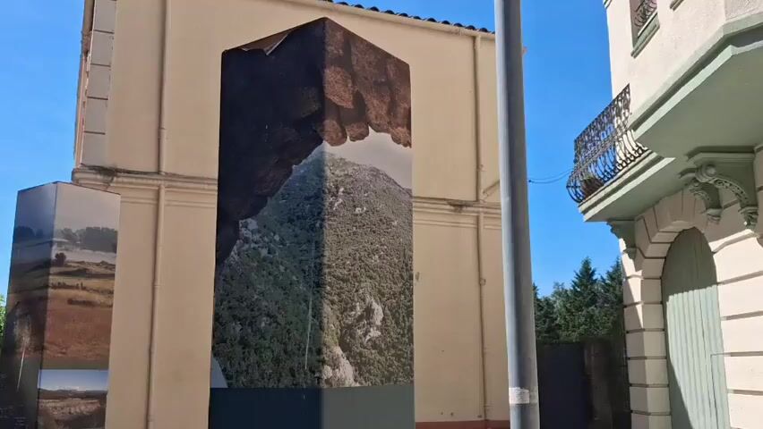 The video shows three dark-colored rectangular pillars on a paved street next to buildings. The pillars have different pictures and text on them. The pillar in the center has a picture of a green mountain underneath a rock overhang, while the pillar to the left has landscape pictures. The camera pans down to show the text on the pillars. The text is in a foreign language. The pillars are labeled with locations: "Canadal," "Santa Llucia," "Sant Julia dels Torts," and "Parissars." A person wearing a white shirt and dark pants walks past the pillars on the street. The weather appears sunny with a blue sky.