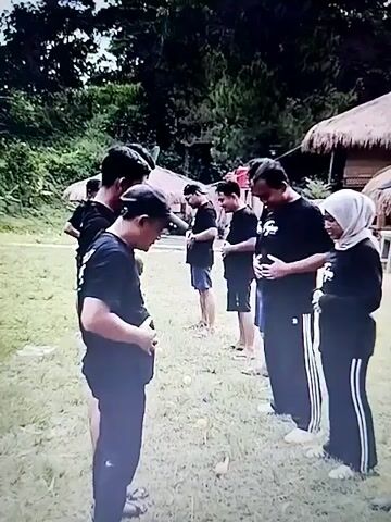 A group of people in black shirts and some in head coverings are outdoors on a grassy area with trees and thatched-roof structures in the background. They appear to be participating in a team-building activity or game. Several individuals are bent over, holding hands, or interacting closely in a playful manner. Some are laughing and smiling, indicating a positive and lighthearted atmosphere. One person is being lifted or is falling, suggesting a physical challenge or activity. The environment is casual and appears to be a recreational or outdoor event.