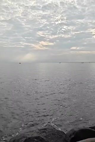 The video shows a view of the sea under a cloudy sky. The water is calm, with small waves creating a gentle ripple effect on the surface. In the distance, several small boats are visible, silhouetted against the horizon. The sky is covered with a pattern of clouds, with some patches of light breaking through. The foreground features a rocky shoreline. The overall scene is serene and peaceful, with a sense of vastness and tranquility.