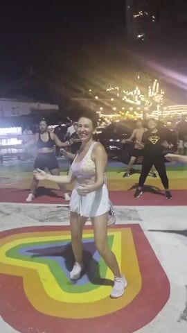 A group of people are dancing outdoors at night. The ground beneath them is painted with a rainbow pattern. A woman in the foreground is wearing a sports bra and white shorts, with white sneakers. She is smiling and appears to be leading the dance. Other people in the background are also dancing. There are lights and buildings visible in the background, suggesting an urban or entertainment district setting.