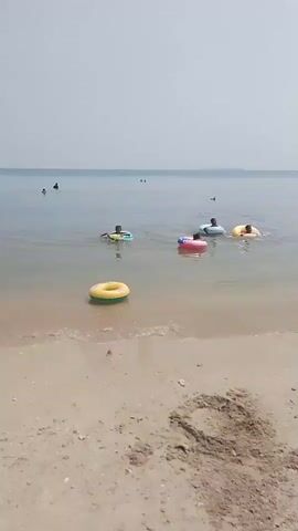 The video shows a sandy beach with calm water. Several people are visible in the water, some using inflatable rings. In the foreground, a yellow inflatable ring with a green stripe is floating near the shore. In the background, more people are swimming or standing in the water. The sky is overcast, and the water appears to be shallow near the beach. The overall scene depicts a relaxing day at the beach.