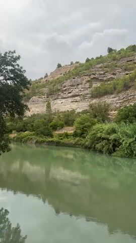 The video shows a scenic view of a river with green water flowing in the foreground. The riverbanks are lined with lush green trees and vegetation. In the background, a rocky hillside rises steeply, dotted with sparse trees and shrubs. The sky is overcast with dramatic clouds, suggesting a moody atmosphere. The water reflects the surrounding greenery and the cloudy sky, creating a peaceful and tranquil scene. There are no people or man-made objects visible in the video, emphasizing the natural beauty of the landscape. The camera pans slightly, offering a broader perspective of the river and the hillside.