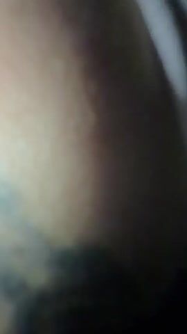 The video shows a person's upper arm with a tattoo that includes a dog's face within a dreamcatcher design. The person is wearing a black jersey with pink and red accents. Another smaller tattoo is visible on the forearm along with some writing. The person appears to be seated or lying down on a gray fabric surface. The lighting is dim.