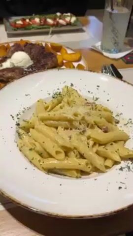 The video shows a table at a restaurant. A plate of penne pasta with a creamy sauce, bacon bits, and parsley is in focus. Behind it, slightly out of focus, is a plate with a grilled steak topped with a dollop of butter, potato wedges, and a small salad. In the background, someone is eating a salad with a fork, and a glass of clear liquid with ice is visible.