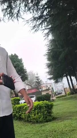 A young man wearing a white collared shirt and black pants stands outdoors under a tree, holding a dark-colored object in his hands. The camera pans to show another young man sitting down and looking at a cell phone. The scene then transitions to a wide shot of a bamboo-like fence structure in front of a tree.