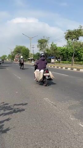 A street view shot shows a person riding a scooter on a paved road. The person is wearing a hooded jacket and a helmet. The scooter is black with a red tail light. The road is divided by a white line. There are trees and streetlights on the side of the road. The sky is blue with some clouds. Another scooter is visible in the background. The time is midday, based on the lighting and shadows.
