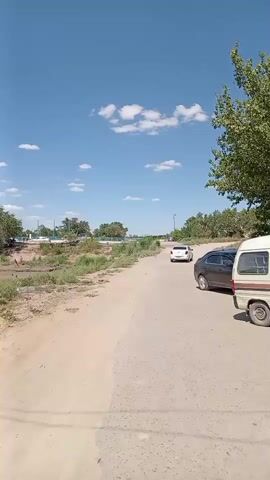The video pans across a dry, dusty landscape under a bright blue sky with scattered white clouds. A dirt road runs diagonally across the frame, bordered on the left by a low embankment with sparse vegetation and what appears to be a small body of water or ditch. On the right side of the road, two cars are visible. The first car, dark in color, is parked on the shoulder. Further ahead, a white car is driving away from the camera. In the distance, across the embankment, there are several trees and some structures, including what might be utility poles and buildings. The overall impression is one of a rural or undeveloped area on a sunny day.