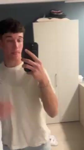 A young man stands in a room, holding a cell phone, taking a selfie in a mirror.  He wears a light-colored t-shirt, light-wash blue jeans, and white sneakers.  He has short, dark brown hair and a bracelet on his left wrist. He gestures with his free hand to his hair and appears to be speaking. In the background is a bed and a white wardrobe.