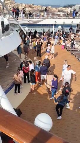 The video shows a high-angle view of the deck of a cruise ship with a crowd of people gathered. The deck is wooden and there are white structures along the perimeter of the ship. People of various ages and ethnicities are present, dressed in casual clothing. Some people are standing still, while others are moving around, seemingly dancing or walking. In the background, a city skyline is visible, suggesting that the ship is near land. The lighting indicates it's daytime, and the overall atmosphere appears relaxed and social.