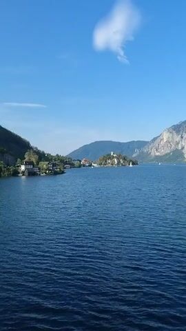 The video captures a panoramic view of a large, calm lake under a clear blue sky with a few wispy white clouds. Mountains, some with rocky, steep faces and others covered in green trees, rise from the far shore. On the left side of the lake, a small island with buildings and trees is visible, with houses and more trees lining the shore to the left of the island. Several sailboats with white sails are scattered across the blue water. The camera pans slowly to the right, revealing more of the mountainous landscape. Towards the end of the video, trees in the foreground partially obscure the view of the lake and mountains on the right side. The overall scene is serene and picturesque, depicting a natural landscape with human elements.