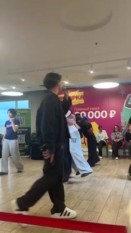 The video captures a lively dance event taking place indoors, likely in a shopping center or event space given the banners and lighting. A group of young people are engaged in a dance battle or performance. The main focus is on the dancers, dressed in casual, streetwear-style clothing. Their movements appear energetic and expressive, suggesting a hip-hop or street dance genre.

In the foreground, a young man in a black jacket and pants is holding a microphone, appearing to be an MC or presenter, interacting with the dancers and the audience. Other individuals are visible in the background, some observing the performance from the side, while others are on the dance floor.

A large banner in the background features Cyrillic text. The most prominent words visible are "ЯРМАРКА" (Yarmarka), meaning "Fair" or "Market," and "Главный приз 30 000 ₽" (Glavnyy priz 30 000 ₽), indicating a grand prize of 30,000 Russian Rubles. This suggests it's a competition or an event with a significant prize. The presence of a "Subway" logo on the banner hints at a possible sponsorship.

The lighting suggests it is daytime, but indoors, so natural light is not a significant factor. The overall atmosphere appears to be exciting and engaging, with the dancers displaying a range of emotions from concentration to joy. The floor is wooden, and the walls are painted in muted tones, with some ceiling lights visible. The presence of a red carpet or demarcation line in the foreground suggests a designated performance area.