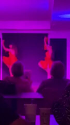 Two women are performing a dance routine on a stage, dressed in purple dresses with ruffles and long gloves. The lighting shifts between blue, pink, and red, creating different moods. An audience of seated people watches the performance. There are tables with cups and mugs in front of them. The room appears to be a lounge or restaurant.