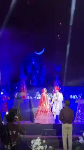 The video captures a wedding celebration at night. A couple, dressed in traditional Indian attire, stands on a stage illuminated by blue lights and flanked by white floral arrangements. Behind them, performers in red costumes add to the festive ambiance. The highlight is a spectacular fireworks display, with golden trails and bursts of light filling the night sky. A photographer and other guests are seen capturing the moment. The atmosphere is vibrant and joyous.