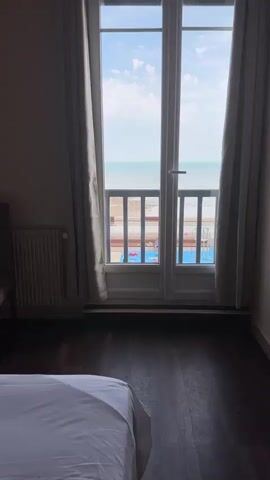 The video opens with a view from a room looking out a double glass door. A white bed with white sheets is visible in the foreground on the left. A white radiator is on the wall to the left of the door. The glass door opens to a view of a railing, a beach, and the ocean under a blue sky with scattered clouds. There is a railing with horizontal bars in front of the door. Beyond the railing, there is a paved area with two blue pools. A person wearing a dark top and shorts is standing near the railing, and a dog is visible below the railing on the left. To the right of the door, the curtain is pulled back, revealing more of the scene. A hand briefly enters the frame from the right, reaching towards the door handle. The video zooms in slightly, providing a closer view of the ocean and the beach area.