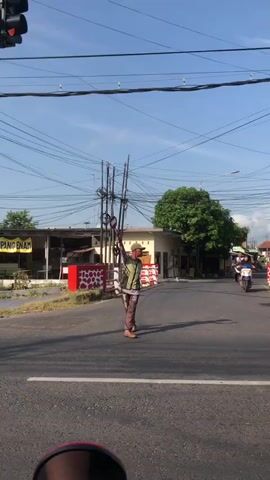 The video captures a street scene with a clear blue sky and overhead power lines. In the foreground, a red and white motorcycle mirror is visible. The camera pans right to show the side of a silver car with tinted windows. Further down the road, a person dressed in light brown pants and a lighter colored shirt, wearing a cap and carrying a long object, stands on the asphalt. They appear to be directing traffic. To the right of this person, a motorcyclist in a red shirt and dark helmet rides past, followed by another motorcycle carrying two people, one in a blue jacket and the other in a dark jacket and cap. The background features buildings, some with red and white decorative elements, and a large green tree. The scene suggests a busy intersection with traffic flow.