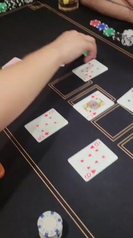 The video shows a close-up of a poker game at a table with a black felt surface. There are several playing cards laid out on the table, along with poker chips in various colors. A person's arm is visible, reaching to place chips on the table. Another person's arm is also visible, holding chips. There's a glass with a drink on the table and a dealer button. The lighting suggests an indoor environment, likely a home or recreational setting. The audio includes a conversation in Russian, discussing betting amounts.