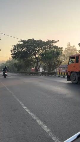 The video shows a street scene with several motorbikes and a truck. The perspective is from the backseat of a motorbike looking forward. In the foreground, the motorbike rider is wearing a black jacket and a helmet. A large truck is visible on the right side of the road, moving in the same direction. A line of motorbikes is proceeding ahead, some single riders and some with passengers. The environment appears to be outdoors on a paved road, with trees and utility poles lining the sides of the street. The sky is clear and bright, suggesting it is daytime, possibly early morning or late afternoon due to the soft lighting. The traffic appears to be moderate, with vehicles moving in the same direction on the road. The overall impression is a typical urban or suburban street scene with vehicular movement.