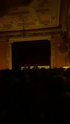 Here's a description of the scene:

The scene is an interior shot of a theater or concert hall. A stage is visible at the front, bordered by ornate golden trim and red walls. The stage is dark, but the silhouettes of several figures can be seen on it. A large audience fills the foreground, their faces mostly obscured by darkness. Overhead, the ceiling has a decorated, patterned design with a chandelier hanging down. The overall lighting is dim, with a yellowish tint.