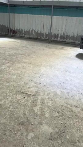 This media file captures a short clip inside a large, open-air warehouse or industrial space in Takfan, Tajikistan. The ground is uneven and dusty, composed of dirt and gravel. The space is characterized by numerous large shipping containers, some stacked and others serving as makeshift structures or offices. Metal walkways and staircases are visible, suggesting an organized but utilitarian environment.

The activity depicted appears to be the movement and arrangement of items within the warehouse. In the first few seconds, a black pickup truck with its driver's side door open is stationary. A person, wearing a red top, is visible inside the truck. Subsequently, a man in dark clothing is seen walking, carrying what appears to be a framed picture or a flat panel, near a blue shipping container with visible Chinese text. Another man is seen near a white shipping container.

The overall atmosphere is one of industrial work or storage. The lighting suggests it is daytime, with natural light filtering into the open-sided structure. There are no overt indications of specific weather conditions, but the dust on the ground implies a dry environment. The visible text on the blue shipping container reads "SIN" and Chinese characters, likely indicating its origin or destination. The scene conveys a sense of ongoing operations within a functional, perhaps developing, industrial area.
