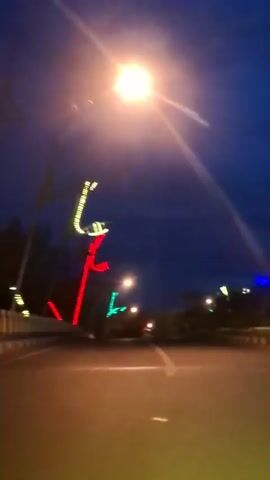 The video shows a dark blue sky over a road at night. There are decorative lights on poles along the side of the road, in red, green, and white. A bright street lamp is visible in the upper part of the frame. The camera is moving forward, suggesting the video was filmed from a vehicle. The road has a white line marking the lane.