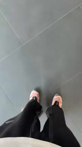 The video starts with a top-down view of someone standing on grey tile flooring, wearing light pink and white sneakers. The camera then pans up to reveal the interior of a car dealership, specifically a Porsche dealership. A white Porsche with a red interior is prominently displayed. There are other vehicles and information kiosks in the background. The camera briefly focuses on the Porsche before returning to the view of the person's feet on the grey tile floor.