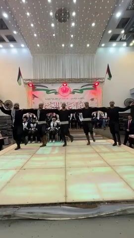 A group of six men, dressed in black clothing and headscarves, perform a synchronized dance on a raised platform. They are holding wooden sticks and their arms are extended, forming a line across the stage. The background features a banner with Arabic writing, Palestinian flags, and red stage lights. Spectators are seated behind the dancers, and the ceiling is decorated with hanging lights.
