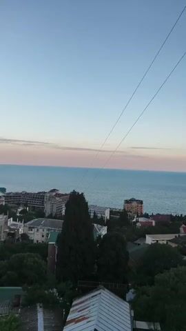 The video is a vertical shot of a coastal town at dusk. Two taut cables run diagonally across the frame from upper left to lower right. Below the cables, the sea stretches to the horizon, with a pastel pink and blue gradient in the sky above it. A shoreline town with various buildings, including apartment complexes and houses, is visible along the coast. Dense green trees are scattered throughout the town and in the foreground. In the lower foreground, the roof of a building with rusty corrugated metal is visible. The scene is peaceful and still.