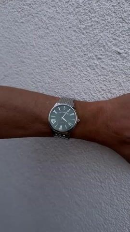 The video begins with a close-up shot of a person's arm wearing a watch against a textured white wall. The watch has a green face with roman numerals and a silver metal band. A second person's arm, also wearing a similar watch, enters the frame from the left. The second person's arm is placed over the first person's arm, creating a cross shape with the wrists. The second person's hand is visible, and they appear to be adjusting or touching the watch on the first person's wrist. The video continues to show the two arms and watches in this crossed position, with slight adjustments in the hand and arm placement. The environment remains consistent throughout the video, with the textured white wall as the background.