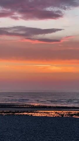 A seascape at sunset. The sky is filled with clouds in shades of pink, orange, and purple. Below the sky, the ocean stretches out, with waves gently lapping the shore. The foreground is a beach covered in dark pebbles. The overall impression is one of tranquility and natural beauty.