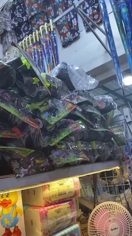 The video shows a stall in a market, likely a toy store or a general store with a toy section. The camera pans across various items, primarily toys, stacked on shelves.

In the foreground, on the lower shelf, there is a stack of toy construction vehicles, predominantly red and yellow dump trucks, packaged in clear plastic. Some of these toys have "TOWN CONSTRUCTION" and "DUMP CONSTRUCTION" labels visible. To the left of these toys, a yellow toy duck with a blue outfit is visible. Next to the dump trucks, there are stacks of pink and white packaged items, possibly tissues or other retail goods. A pink electric fan is partially visible in the bottom right corner.

Above the construction toys, the shelves are filled with more toys, some of which appear to be smaller vehicles like cars or trucks, also in plastic packaging with colorful labels. The packaging of some toys features images of racing cars and characters, suggesting they are aimed at children.

In the background, hanging items are visible. These appear to be clothing, such as t-shirts and pajamas, with various patterns, including floral designs. Several colorful umbrellas are also displayed hanging in the background. The overall environment suggests an indoor market or a shop with a wide assortment of goods. The lighting appears to be artificial, likely from fluorescent lights illuminating the shelves. There are no people clearly visible interacting with the items in the video, indicating it might be a self-service area or the store is not actively attended at the moment of filming.