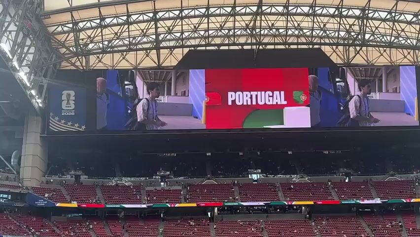 The video shows a wide shot of a soccer stadium. A large jumbotron screen displays content for the FIFA World Cup 2026. On the left side of the screen, the FIFA World Cup 2026 logo is visible. The center and right portions of the screen show visuals related to Portugal. Initially, the word PORTUGAL is displayed in large white letters on a red background, with images of two men walking on either side. Then, the screen changes to show the Portuguese flag and the word PORTUGAL on a green and red background, followed by an animation of the word PORTUGAL repeating. The screen then transitions to show blue team buses with the Portuguese flag and the word PORTUGAL on them, with people standing next to the buses. The stadium seating, mostly empty, is visible in the foreground, with red seats and some green elements. The stadium roof structure is also visible.