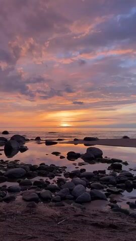 The video shows a tranquil beach scene at sunset.  The sky is awash with hues of orange, pink, and purple, with clouds scattered across the horizon.  The sun is setting over the water, casting a golden glow.  In the foreground, smooth, dark stones of varying sizes are partially submerged in shallow pools of water, reflecting the colors of the sky.  The beach sand is a reddish-brown color. A voice can be heard in the background.