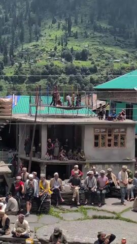 A crowd of people is gathered in what appears to be a village square in a mountainous region. In the foreground, several men are seated on stone benches, some wearing traditional clothing and hats. Behind them, a building with multiple levels is visible. On the rooftop of this building, a group of individuals, some appearing to be performing or engaged in some activity, are standing around a structure with colorful poles. Further back, the hillside is covered with lush green vegetation and scattered buildings, suggesting a rural setting. The overall atmosphere seems to be one of a community gathering or celebration.