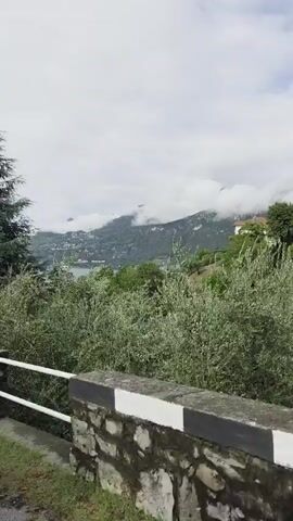 The video pans to the left showing a view of mountains and a village in the distance. In the foreground, there is a stone wall with a metal railing. A large tree with green foliage is visible on the right side of the frame. The sky is cloudy.