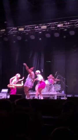 The video shows a group of women performing on a stage. In the foreground, a crowd of people is visible, mostly in silhouette. The stage is lit with bright spotlights, and a dark curtain serves as a backdrop. On stage, four women are visible. One woman in a pink outfit is playing a saxophone. Another woman, wearing a white top and a short pink skirt, is singing into a microphone and dancing. Two other women in pink outfits are also on stage, one of whom appears to be dancing or interacting with the singer. A drum set is visible on a platform behind the performers. The overall atmosphere appears to be a live music performance.