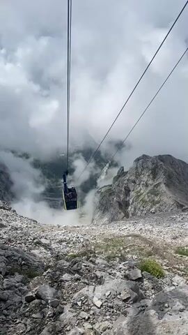 A yellow cable car descends a steep mountain slope. The cable car is suspended from two thick cables, disappearing into the distance. Below the cable car, the mountain drops away into a foggy valley. The ground in the foreground is covered in grey rocks and sparse green vegetation. The sky is overcast with white clouds. A person is visible inside the cable car.