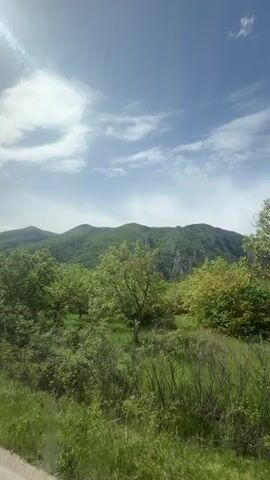 The video shows a landscape of rolling green hills and lush trees under a blue sky dotted with white clouds. The scene is filmed from a moving vehicle, as evidenced by the passing scenery. A black car briefly enters the frame from the lower left. The overall impression is one of natural beauty and a sunny day.