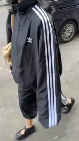 A woman wearing a black Adidas tracksuit and black Mary Jane style shoes walks on a sidewalk, carrying a beige shoulder bag. A car and trash cans are visible in the background.