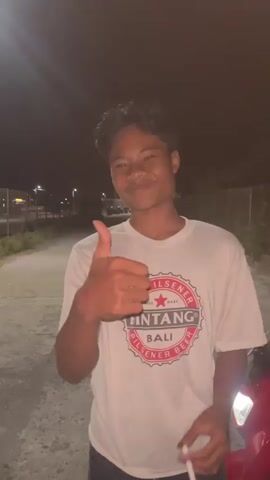 The video shows a group of young people at night. The first frame shows a young person in a white t-shirt with a FINTANG BALI logo, giving a thumbs up to the camera. The background is a dark road with a streetlight. The next few frames show other young people, some interacting with each other and some walking away from the camera. There are trees and fences visible in the background, and the scene appears to be outdoors. The lighting is dim, suggesting it is nighttime.
