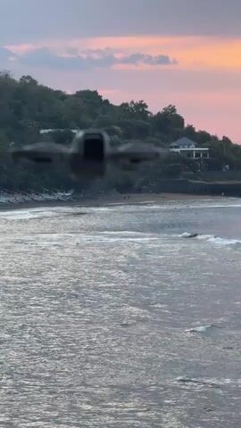 A drone flies over the ocean in front of a beach with a hill and houses in the background. The sky is pink and orange from a sunset.