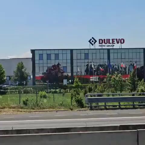 The camera is pointed toward a building complex, apparently seen from a moving vehicle alongside a road with guardrails. The building has a modern design with large glass windows. Above the windows is the name "DULEVO" and "FAYAT GROUP". Several flags are visible in front of the building.  There are a few trucks parked in front of the building. The surrounding environment includes green grass, small trees, and a fence that lines the property.  Other vehicles can be seen moving on the road. The sky is clear and blue.
