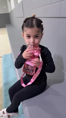 A young girl with dark hair styled in two buns and two pigtails is sitting on a grey bench, holding a pink water bottle with a straw. She is wearing a black long-sleeved shirt and black leggings with white socks that have brown polka dots. The girl is looking directly at the camera with a playful expression, and she appears to be drinking from the bottle. The water bottle has a decorative pink strap with various sea-themed charms, including shells and starfish. A small Russian flag patch is visible on the sleeve of her shirt. The setting appears to be an indoor space, possibly a gymnasium or a recreational facility, with a grey brick wall in the background and a blue mat on the floor to the left. The lighting suggests it is daytime. The girl's expression and the way she is holding the bottle convey a sense of enjoyment. There is some faint text on the water bottle that is difficult to read clearly.