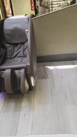 A man in a light-colored t-shirt and dark pants holds up a phone to take a picture or video. He stands near a row of dark brown massage chairs, with the back of one chair facing the camera. The chairs are positioned on a light wood-grain floor in what appears to be a shopping mall. In the background, escalators and store displays are visible. The massage chairs have textured upholstery and illuminated blue accents on their bases.