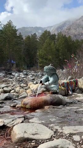 The image displays an outdoor scene centered on a unique fountain featuring a statue. The main subject is a greenish-blue patinated statue, likely made of bronze or a similar weathered metal, depicting a young person or child in traditional attire. The figure is shown kneeling or crouching, wearing a large, fur-lined hat and a thick, quilted-looking jacket. It holds a cylindrical vessel or scoop from which a clear stream of water continuously flows.

The water pours into an irregularly shaped basin, carved from a rough, natural stone, which exhibits a reddish-brown coloration, suggestive of mineral deposits, possibly iron. The ground surrounding the fountain is rugged, covered extensively with various-sized grey and tan rocks, indicating a natural, possibly riverine or mountainous environment.

In the background, a dense forest of tall, green coniferous trees (likely pines or firs) extends upward, with some bare, leafless deciduous branches visible, suggesting an early spring or late autumn setting. The scene is brightly illuminated by natural sunlight, creating clear visuals and subtle shadows. The overall atmosphere is tranquil, highlighting a natural spring or mineral water source, common in areas like Arshan, Russia, known for its therapeutic waters. No visible text is present in the image.