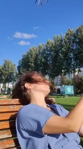 A young woman is sitting on a park bench outdoors on a sunny day. She is wearing a blue top and has long brown hair. Her head is tilted back, and her eyes are closed. A blue kite with yellow and red streamers is flying in the clear blue sky above her. In the background, there are trees and some buildings. The woman is holding the string of the kite, which is taut as it flies in the wind.