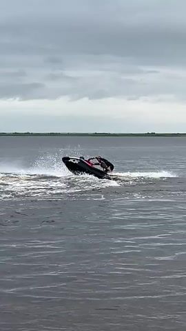 A jet ski is performing a stunt on the water. A person is standing on the back of the jet ski, leaning over and holding onto the handlebars. The jet ski is tilted up on its side, with a large wake of water behind it. The sky is cloudy, and there is a body of water stretching to the horizon.