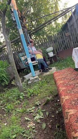 A woman is sitting on a swing set and holding a small child who is standing in front of her. The woman is wearing a white and blue patterned shirt and dark pants. The child is wearing a yellow shirt and dark pants. The swing set is made of metal and is painted blue and white. The background shows a wooden fence, trees, and grass. There is also a rug or blanket draped over a surface in the foreground. The scene appears to be in a backyard or outdoor area.