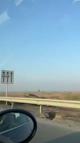 The video shows a view from inside a car, likely the driver's perspective, as it moves along a road adjacent to a dry, barren landscape. The sky is clear and blue, though there's some haze visible in the distance. The landscape is mostly flat with sparse, low-lying vegetation. A side mirror is visible in the foreground. No people are clearly visible. The car is moving forward.